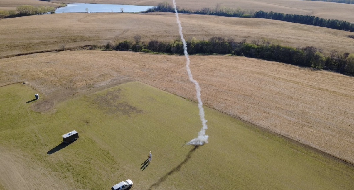 Aerial view of a model rocket launch: white smoke trail above an open field, with vehicles and range setup visible.